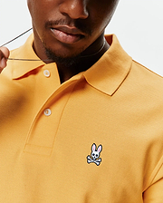Mens Classic Pique Polo in mustard yellow, featuring a classic fit, textured pique fabric, white button placket, and a small embroidered white and pink Psycho Bunny skull logo on the left chest.
