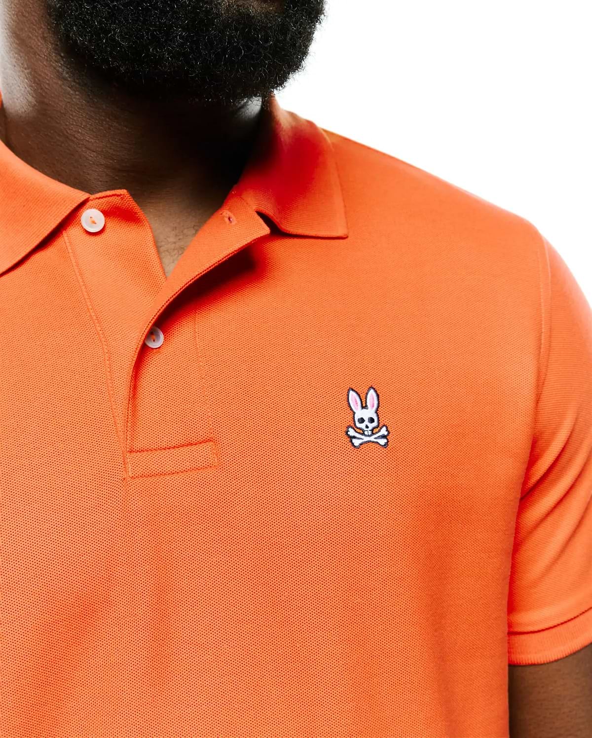 Men's Classic Pima Pique Polo in vibrant orange pique knit, featuring a small embroidered white and pink Psycho Bunny skull logo on the left chest and a two-button placket with white buttons. (2)