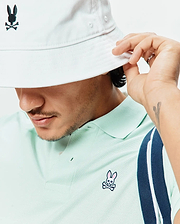 Mens Classic Pique Polo in pale mint green pique, classic fit, with an embroidered pink, black, white Psycho Bunny logo on the left chest and a navy/white striped sleeve. White bucket hat with black Psycho Bunny logo. (3)