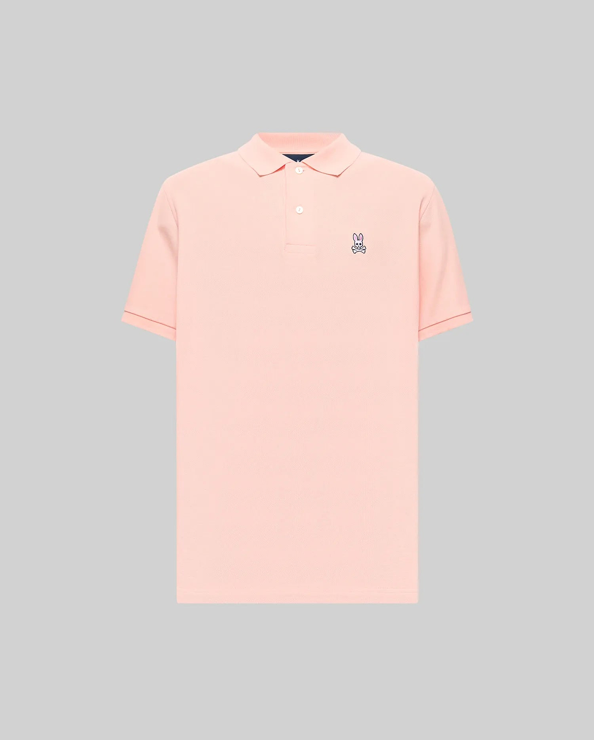Mens Classic Pique Polo in peach, featuring a classic fit, pique knit texture, two-button placket, and a small embroidered purple, white, and black Psycho Bunny skull logo on the left chest with subtle red cuff tipping.