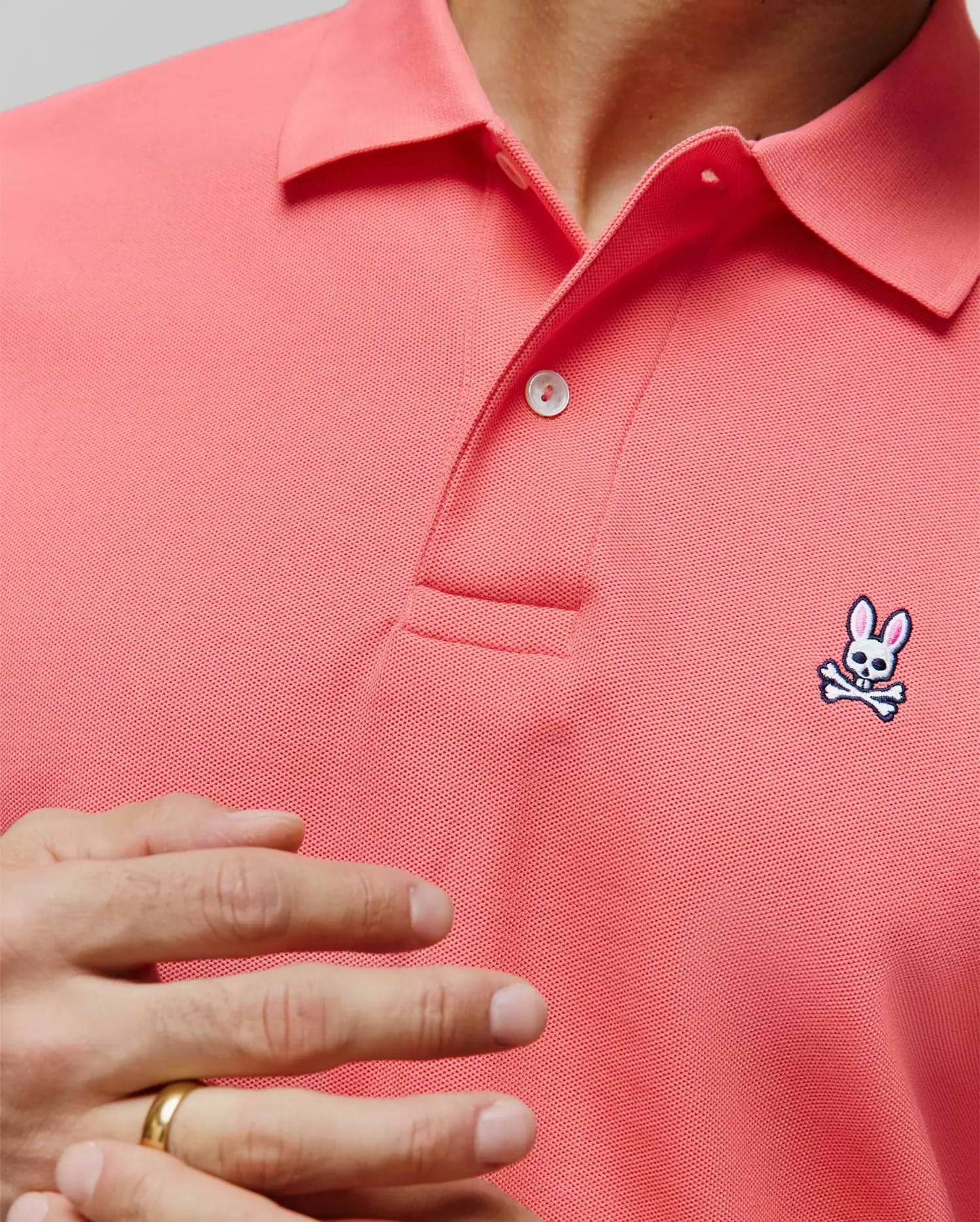 MENS CLASSIC PIQUE POLO in vibrant coral, featuring a classic fit, textured pique fabric, two-button placket, and a small embroidered Psycho Bunny skull logo on the left chest with white, black, and pink accents.