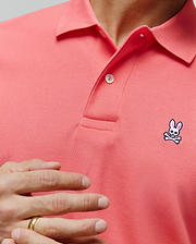 MENS CLASSIC PIQUE POLO in vibrant coral, featuring a classic fit, textured pique fabric, two-button placket, and a small embroidered Psycho Bunny skull logo on the left chest with white, black, and pink accents.