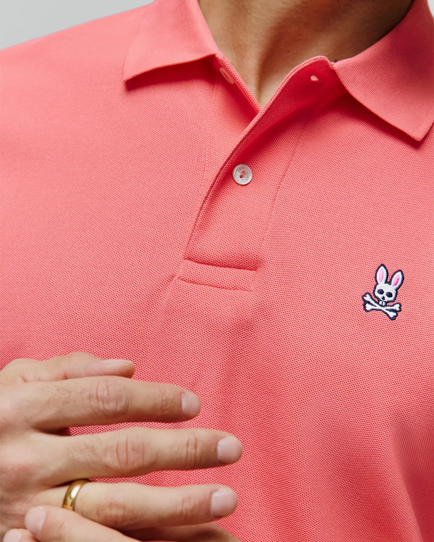 MENS CLASSIC PIQUE POLO in vibrant coral, featuring a classic fit, textured pique fabric, two-button placket, and a small embroidered Psycho Bunny skull logo on the left chest with white, black, and pink accents.
