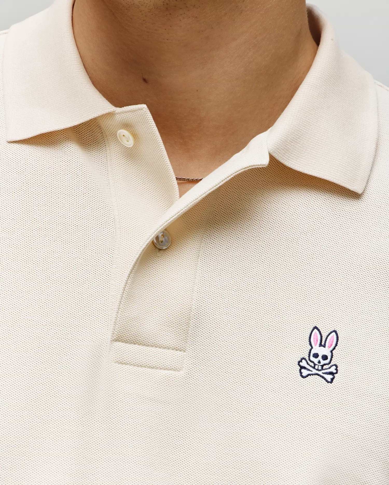 Close-up of a person wearing the Psycho Bunny MENS CLASSIC PIQUE POLO SHIRT - B6K001F200 in cream, with two buttons undone and a small embroidered Bunny logo with pink ears and crossbones on the left chest.