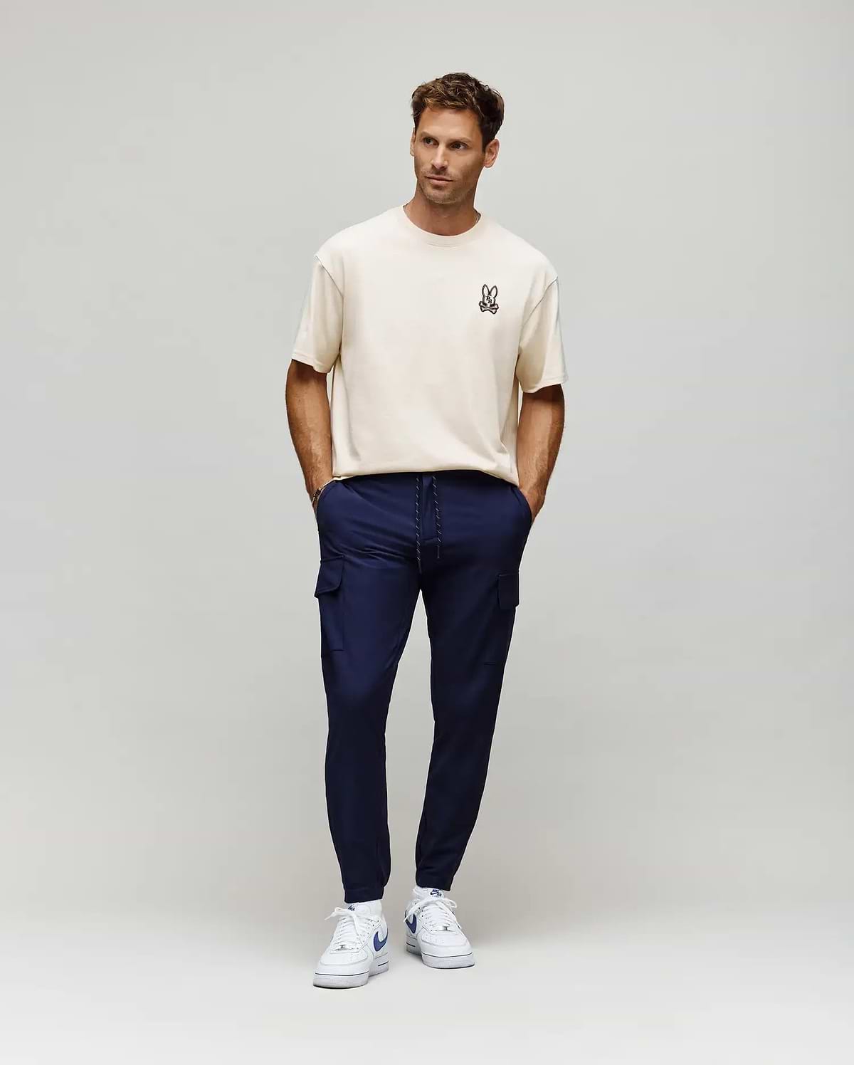 Ecru relaxed fit jersey t-shirt with a small embroidered Psycho Bunny skull logo on the left chest, paired with slim navy Mens Lloyd Cargo Jogger pants featuring cargo pockets and a white drawstring.