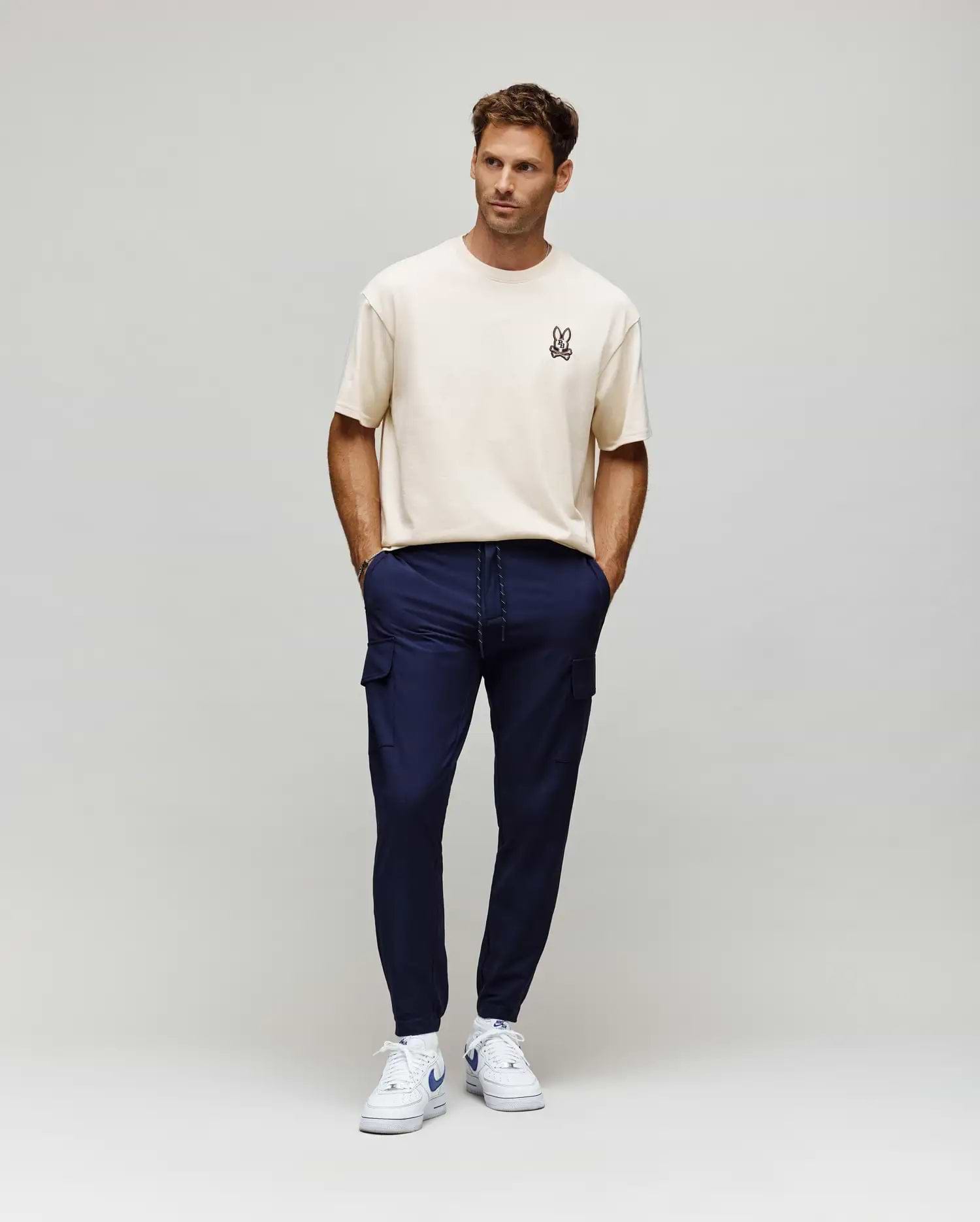 Ecru relaxed fit jersey t-shirt with a small embroidered Psycho Bunny skull logo on the left chest, paired with slim navy Mens Lloyd Cargo Jogger pants featuring cargo pockets and a white drawstring.
