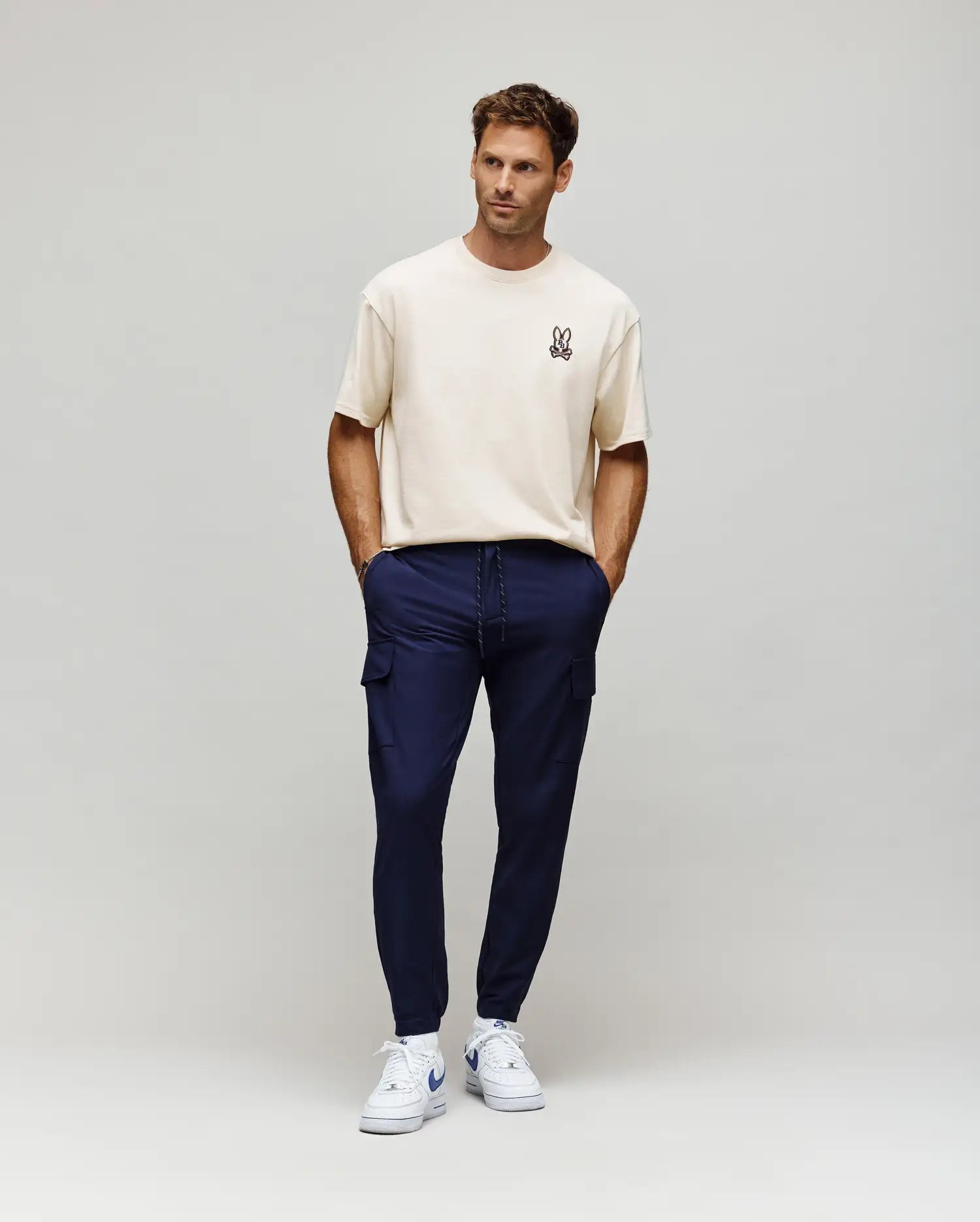 Ecru relaxed fit jersey t-shirt with a small embroidered Psycho Bunny skull logo on the left chest, paired with slim navy Mens Lloyd Cargo Jogger pants featuring cargo pockets and a white drawstring.