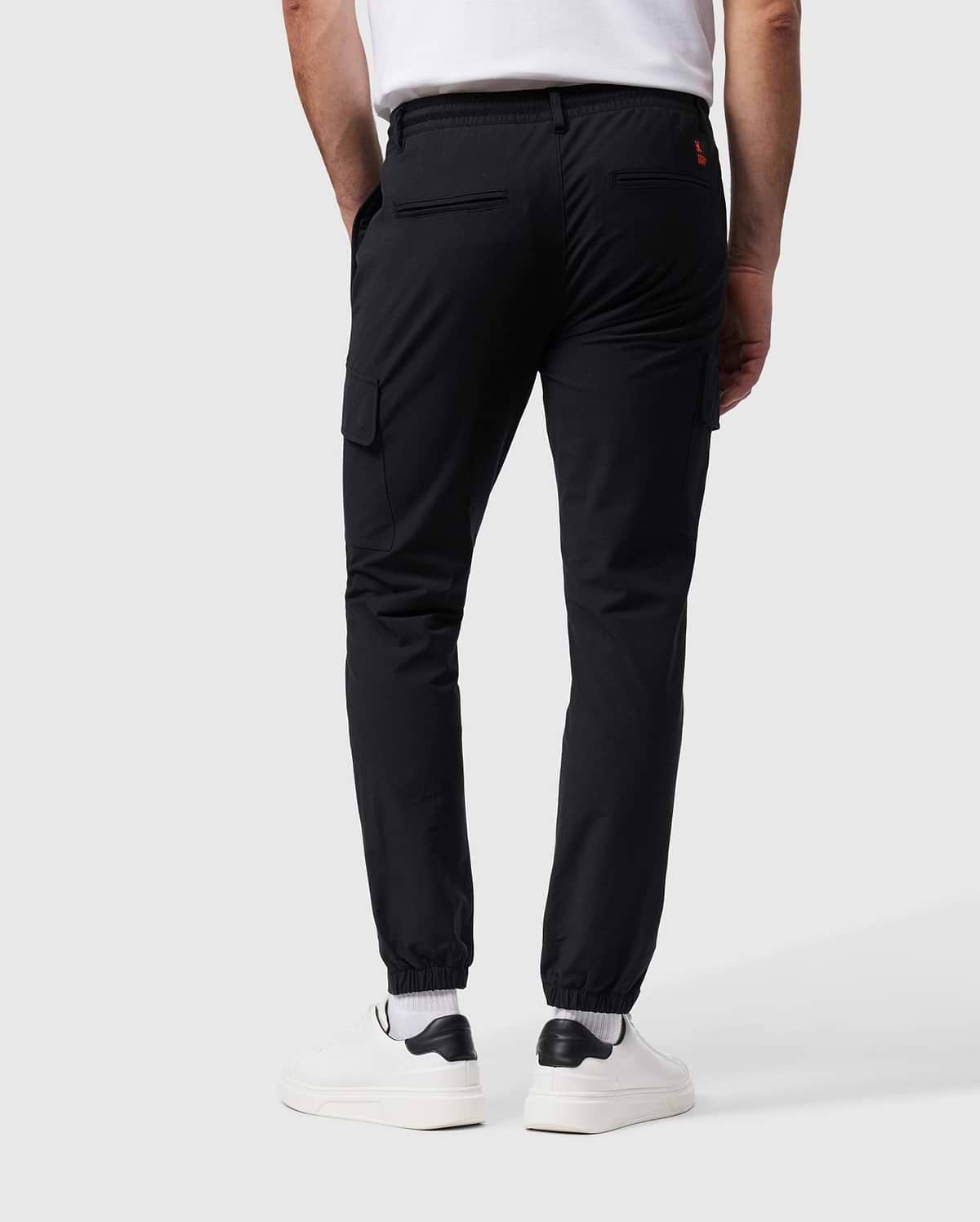 A person wearing Psycho Bunny's MENS LLOYD CARGO JOGGER - B6P159B200 in black, featuring elasticized cuffs and utilitarian pockets, paired with a white T-shirt, white socks, and white sneakers. The person is standing with their back facing the camera against a plain gray background.