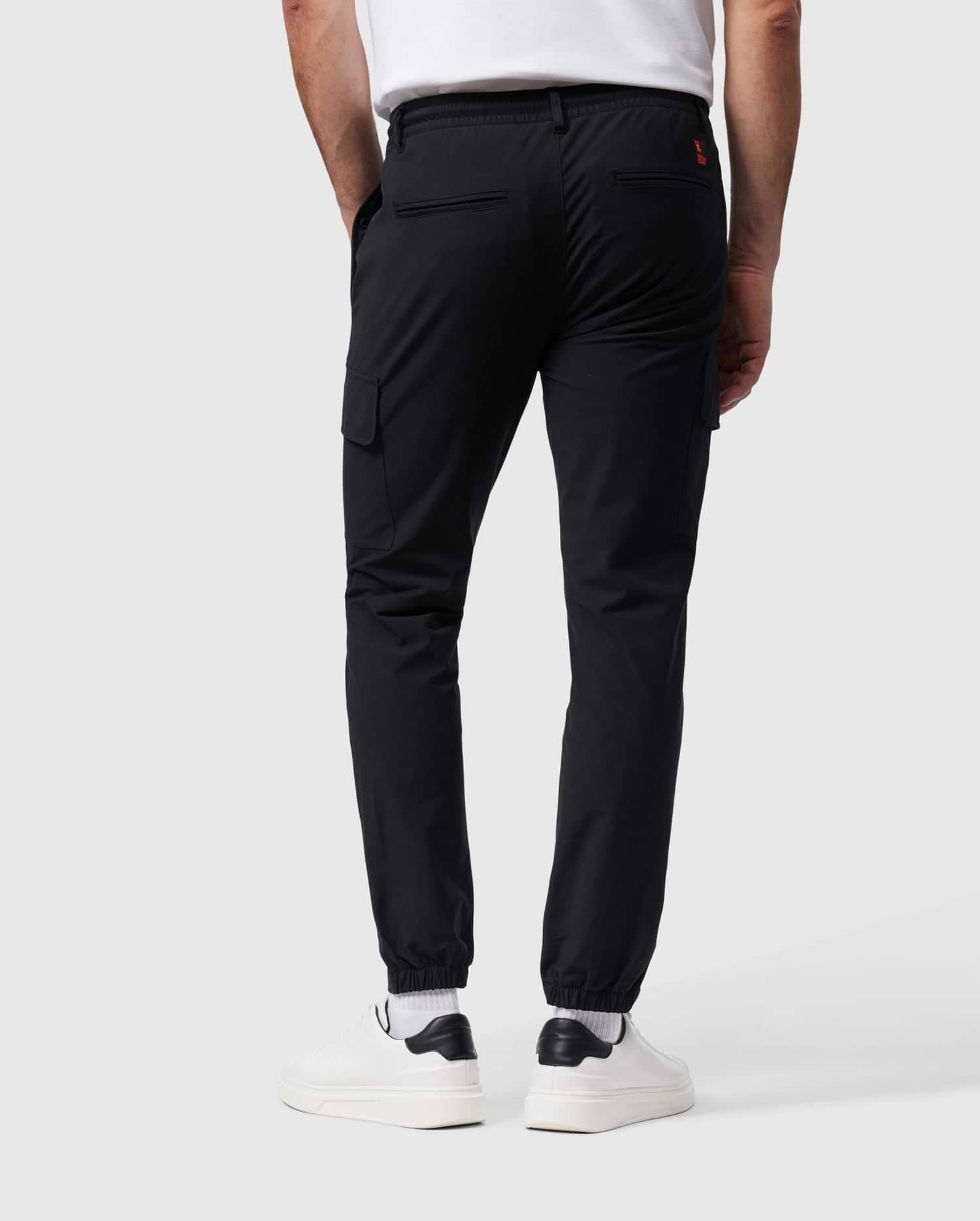 A person wearing Psycho Bunny's MENS LLOYD CARGO JOGGER - B6P159B200 in black, featuring elasticized cuffs and utilitarian pockets, paired with a white T-shirt, white socks, and white sneakers. The person is standing with their back facing the camera against a plain gray background.