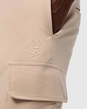 Close-up of a pocket on the MENS LLOYD CARGO JOGGER - B6P159B200 by Psycho Bunny, showcasing a stitched logo of a bunny head with crossbones underneath. The smooth fabric pocket has a flap secured by snap fastening, and a hand is casually resting in the side pocket, highlighting the refined embroidery.