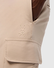 Close-up of a pocket on the MENS LLOYD CARGO JOGGER - B6P159B200 by Psycho Bunny, showcasing a stitched logo of a bunny head with crossbones underneath. The smooth fabric pocket has a flap secured by snap fastening, and a hand is casually resting in the side pocket, highlighting the refined embroidery.