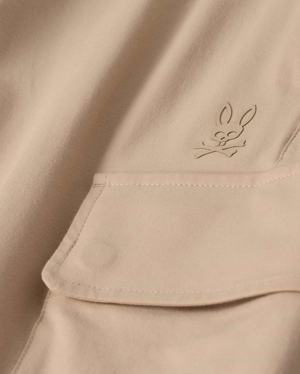 Close-up of the MENS LLOYD CARGO JOGGER - B6P159B200 by Psycho Bunny, featuring a beige fabric adorned with a snap fastening pocket. Above the pocket is a stitched bunny logo with crossed bones underneath its head, showcasing a subtle pirate motif. The playful embroidery refinement adds an elegant touch to the smooth and well-crafted fabric.