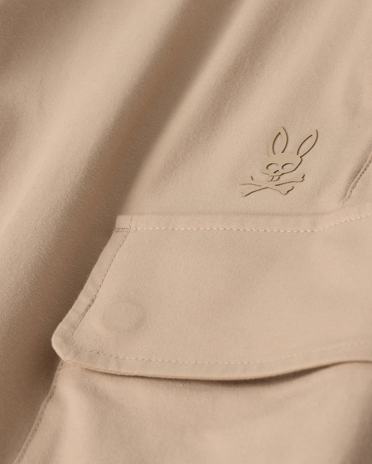 Close-up of the MENS LLOYD CARGO JOGGER - B6P159B200 by Psycho Bunny, featuring a beige fabric adorned with a snap fastening pocket. Above the pocket is a stitched bunny logo with crossed bones underneath its head, showcasing a subtle pirate motif. The playful embroidery refinement adds an elegant touch to the smooth and well-crafted fabric.