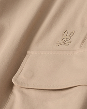 Close-up of the MENS LLOYD CARGO JOGGER - B6P159B200 by Psycho Bunny, featuring a beige fabric adorned with a snap fastening pocket. Above the pocket is a stitched bunny logo with crossed bones underneath its head, showcasing a subtle pirate motif. The playful embroidery refinement adds an elegant touch to the smooth and well-crafted fabric.