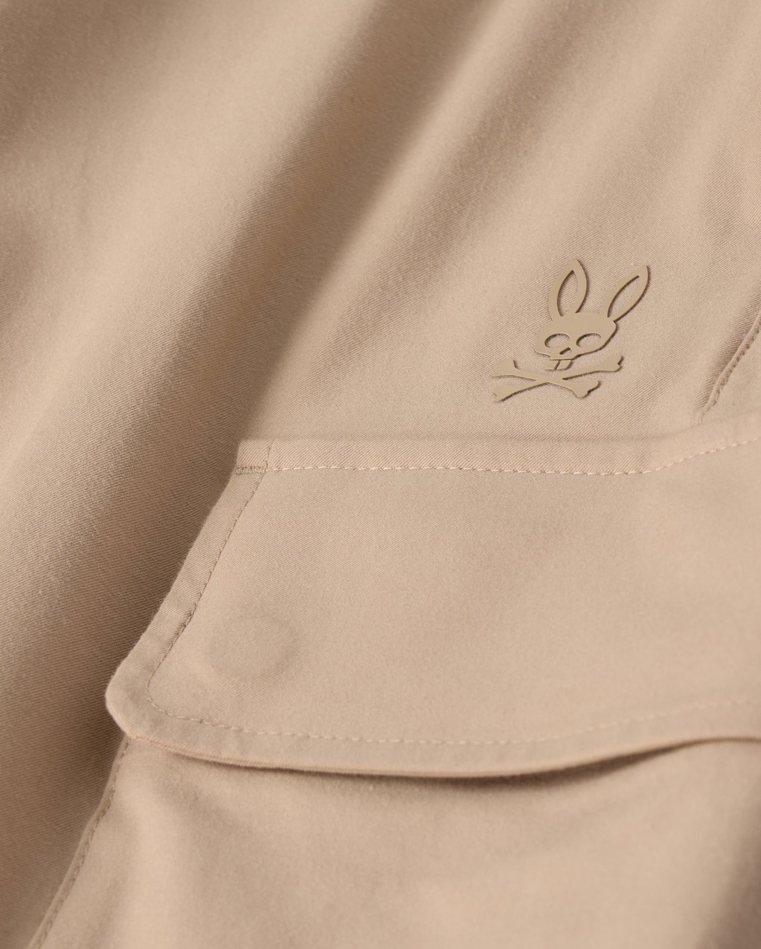 Close-up of the MENS LLOYD CARGO JOGGER - B6P159B200 by Psycho Bunny, featuring a beige fabric adorned with a snap fastening pocket. Above the pocket is a stitched bunny logo with crossed bones underneath its head, showcasing a subtle pirate motif. The playful embroidery refinement adds an elegant touch to the smooth and well-crafted fabric.