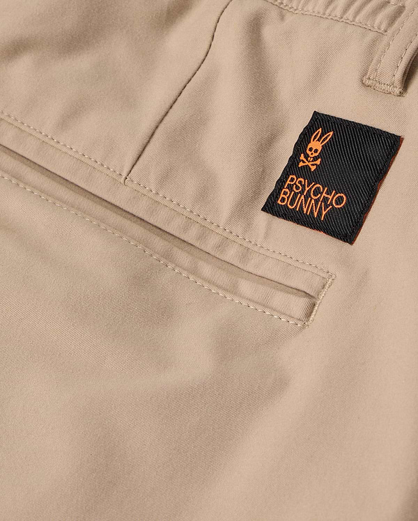 Close-up of the beige fabric MENS LLOYD CARGO JOGGER - B6P159B200 by Psycho Bunny, showcasing an orange-accented black label with a bunny logo and the words "Psycho Bunny" stitched on. The jogger pants exhibit detailed stitching and refined embroidery around the snap fastening pockets.