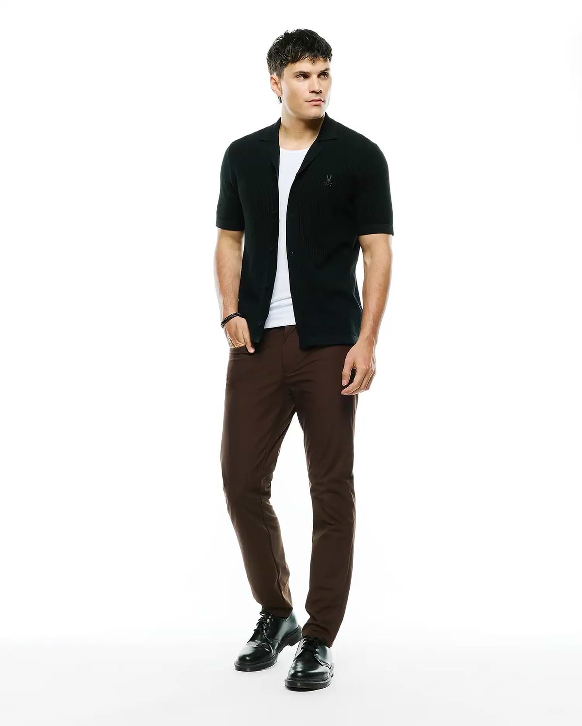 Black ribbed knit short-sleeve collared shirt with embroidered Psycho Bunny skull logo on chest, layered over a white tee, paired with slim-fit brown Mens Maverick 10k Step Pant.