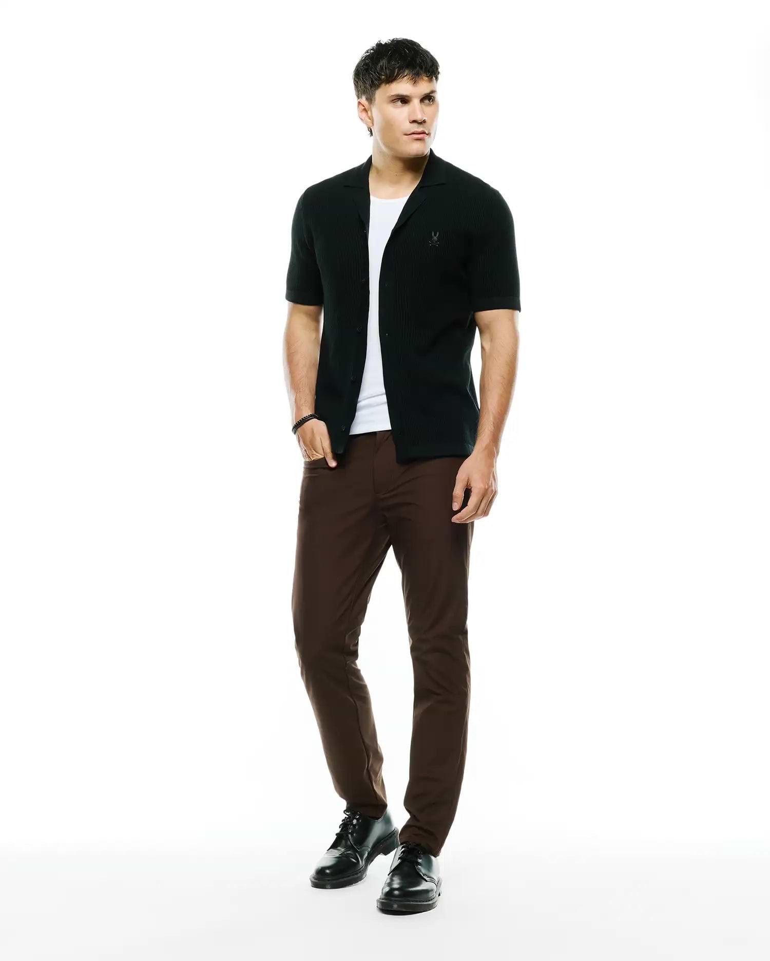 Black ribbed knit short-sleeve collared shirt with embroidered Psycho Bunny skull logo on chest, layered over a white tee, paired with slim-fit brown Mens Maverick 10k Step Pant.