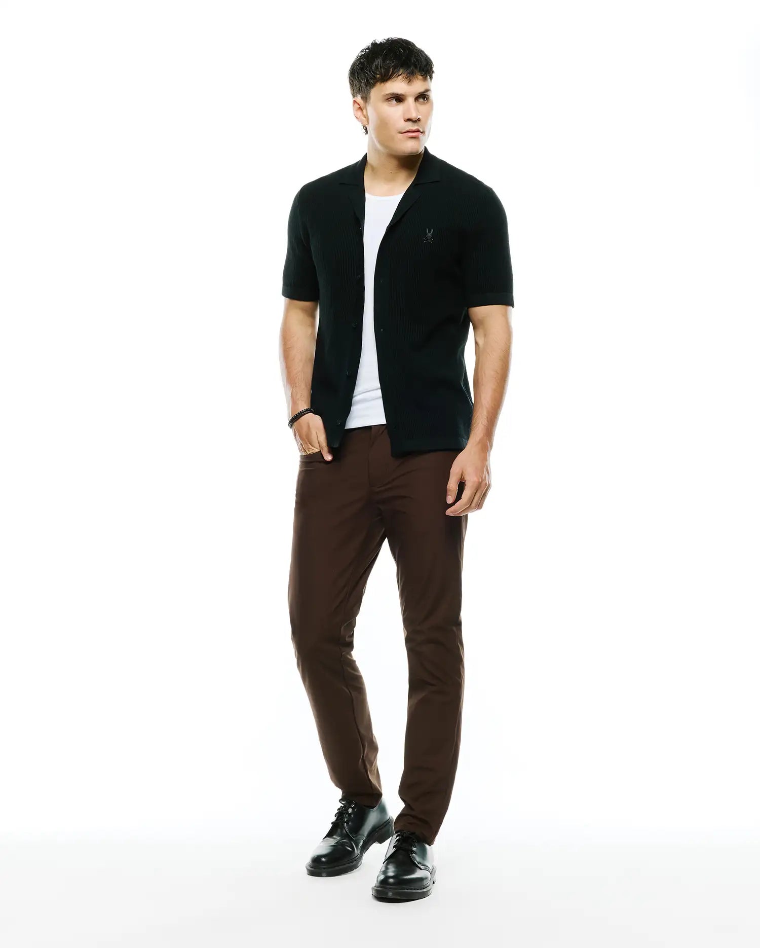 Black ribbed knit short-sleeve collared shirt with embroidered Psycho Bunny skull logo on chest, layered over a white tee, paired with slim-fit brown Mens Maverick 10k Step Pant.