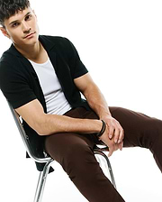 Dark green ribbed knit short-sleeve cardigan with collar and small embroidered Psycho Bunny logo on left chest, layered over a white crewneck t-shirt, paired with slim brown MENS MAVERICK 10K STEP PANT.