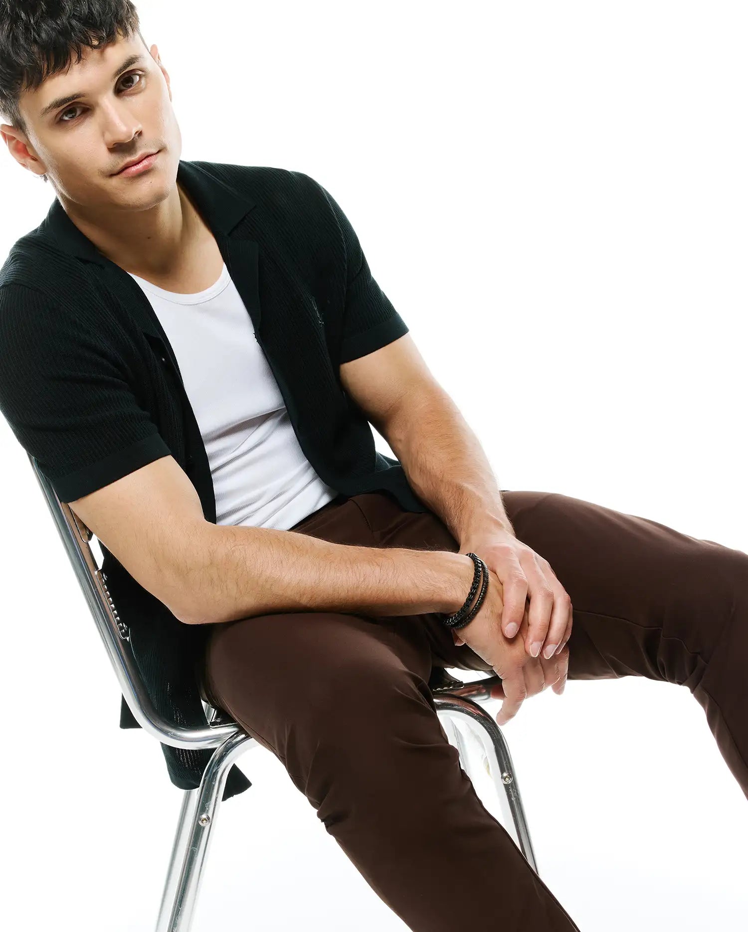 Dark green ribbed knit short-sleeve cardigan with collar and small embroidered Psycho Bunny logo on left chest, layered over a white crewneck t-shirt, paired with slim brown MENS MAVERICK 10K STEP PANT.