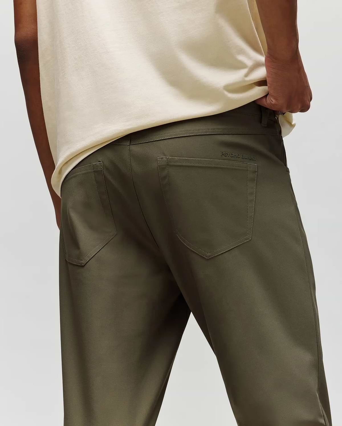 A person is shown from the back wearing Psycho Bunny’s MENS MAVERICK 10K STEP PANT - B6P788D200 in olive-green, with five-pocket styling, a cream shirt, one hand in their pocket, and subtle branding above the right pocket.