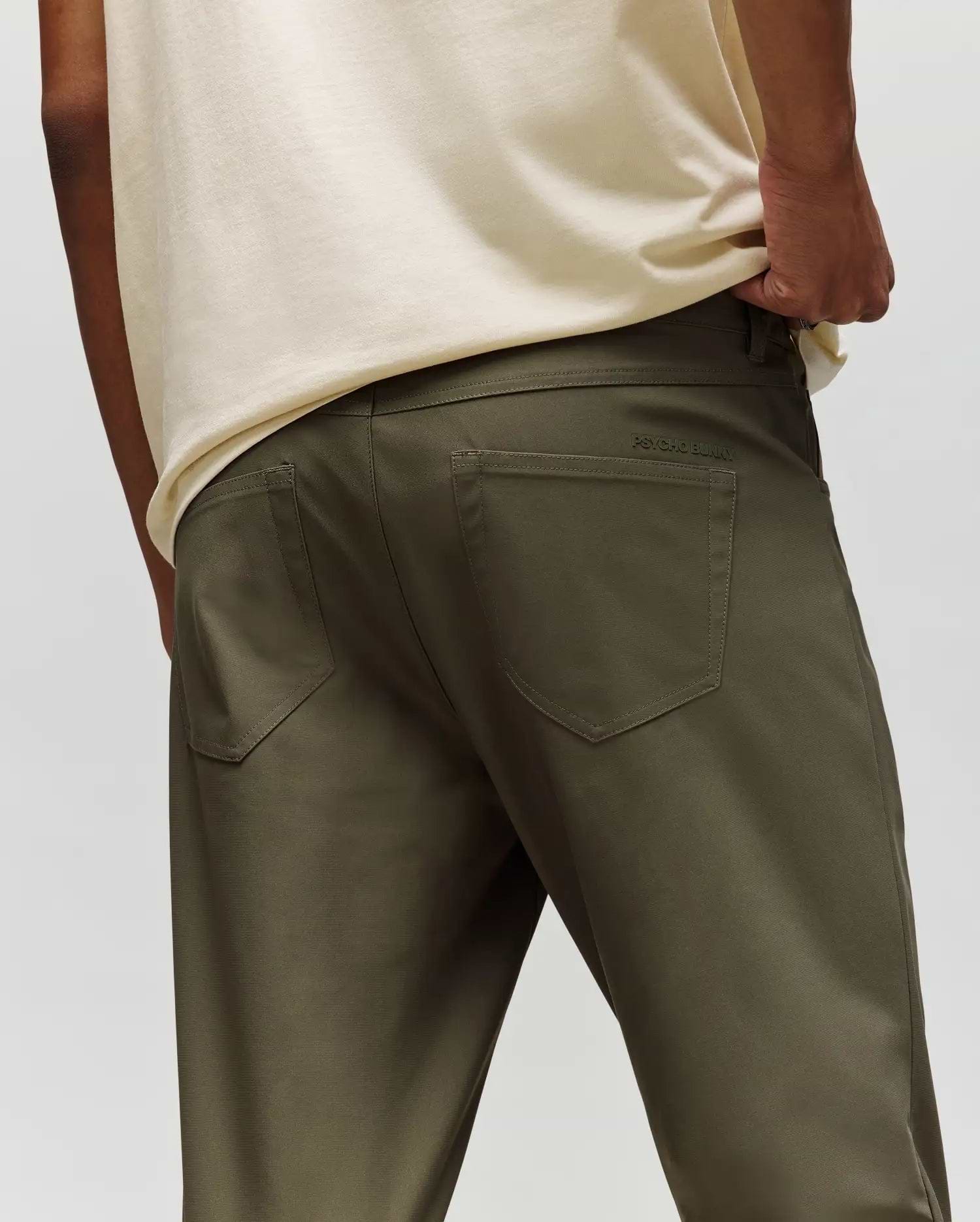 A person is shown from the back wearing Psycho Bunny’s MENS MAVERICK 10K STEP PANT - B6P788D200 in olive-green, with five-pocket styling, a cream shirt, one hand in their pocket, and subtle branding above the right pocket.