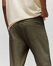 A person is shown from the back wearing Psycho Bunny’s MENS MAVERICK 10K STEP PANT - B6P788D200 in olive-green, with five-pocket styling, a cream shirt, one hand in their pocket, and subtle branding above the right pocket.