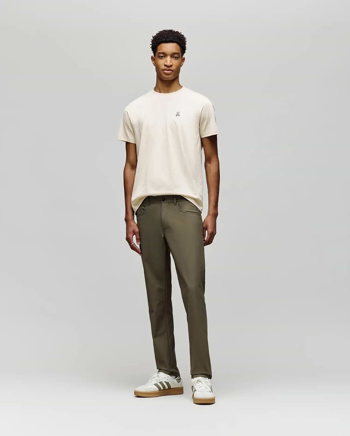 A young man stands against a plain background, wearing a beige t-shirt and Psycho Bunny MENS MAVERICK 10K STEP PANT - B6P788D200. He pairs this with white striped sneakers, facing forward with a neutral expression and relaxed posture.