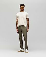 A young man stands against a plain background, wearing a beige t-shirt and Psycho Bunny MENS MAVERICK 10K STEP PANT - B6P788D200. He pairs this with white striped sneakers, facing forward with a neutral expression and relaxed posture.