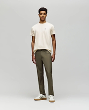 A young man stands against a plain background, wearing a beige t-shirt and Psycho Bunny MENS MAVERICK 10K STEP PANT - B6P788D200. He pairs this with white striped sneakers, facing forward with a neutral expression and relaxed posture.