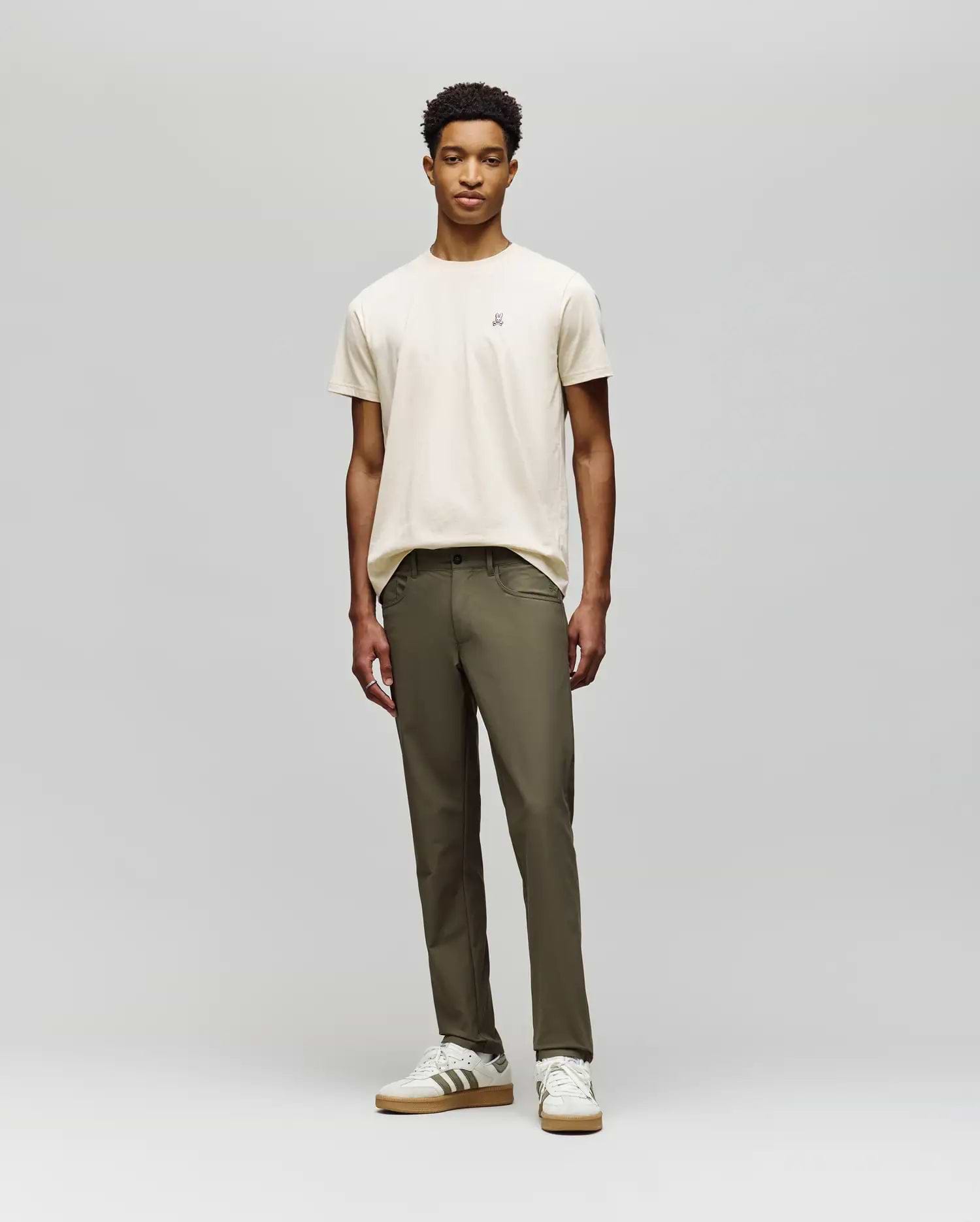 A young man stands against a plain background, wearing a beige t-shirt and Psycho Bunny MENS MAVERICK 10K STEP PANT - B6P788D200. He pairs this with white striped sneakers, facing forward with a neutral expression and relaxed posture.
