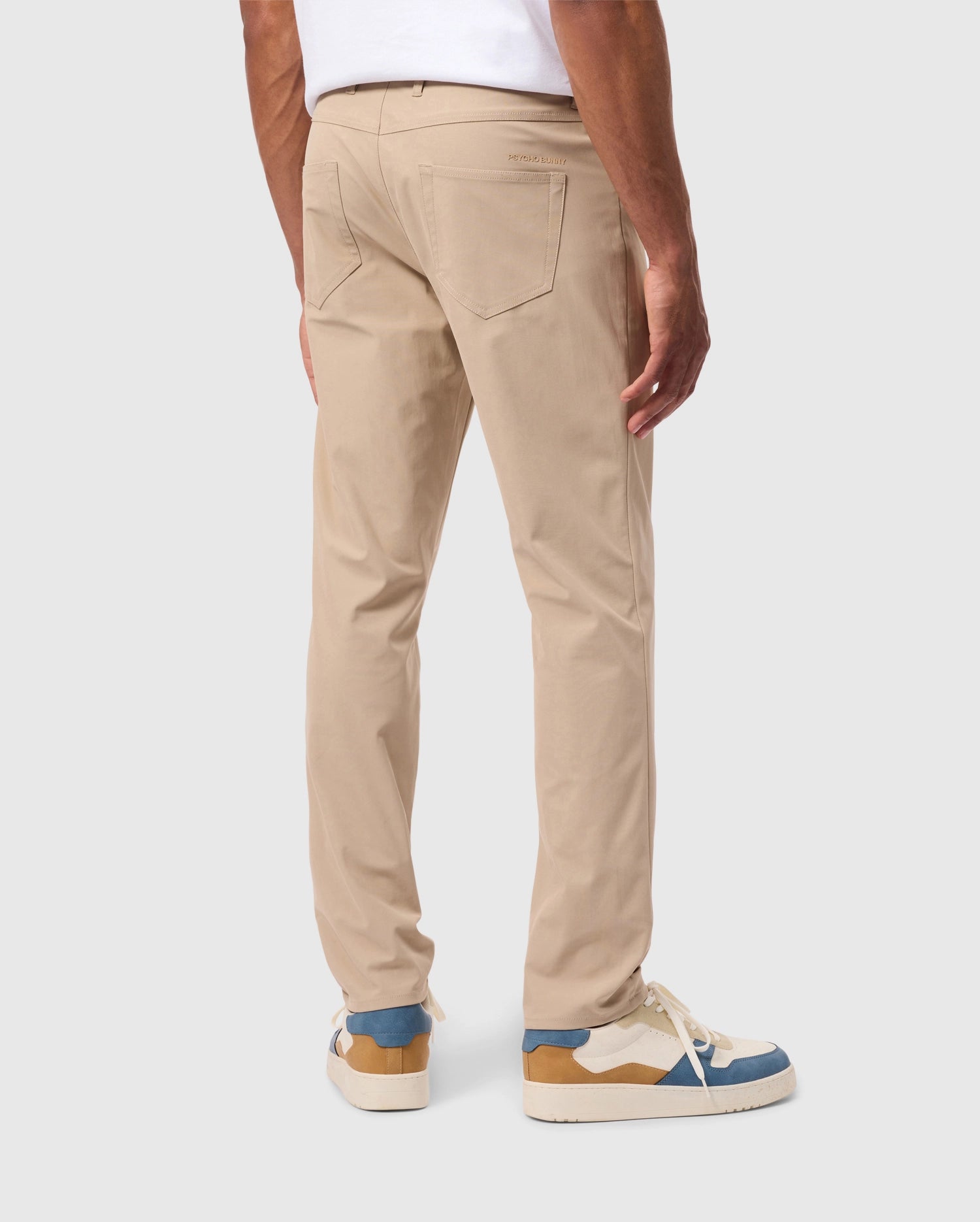 A person is standing, facing away from the camera, dressed in Psycho Bunny's beige MENS 10K STEP PANT (B6P788D200) with a five-pocket design and a white shirt. Their footwear consists of white sneakers with blue and brown accents. The background is plain and light-colored.