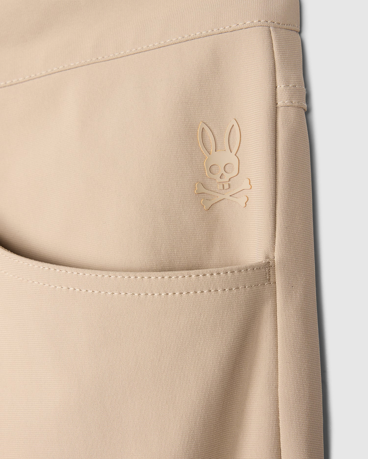 Close-up of a beige pocket on the MENS 10K STEP PANT - B6P788D200 by Psycho Bunny, featuring a raised emblem of a bunny skull and crossbones design. The stitching and fabric texture are visible, showcasing its four-way stretch material. The background is plain white, highlighting the details.