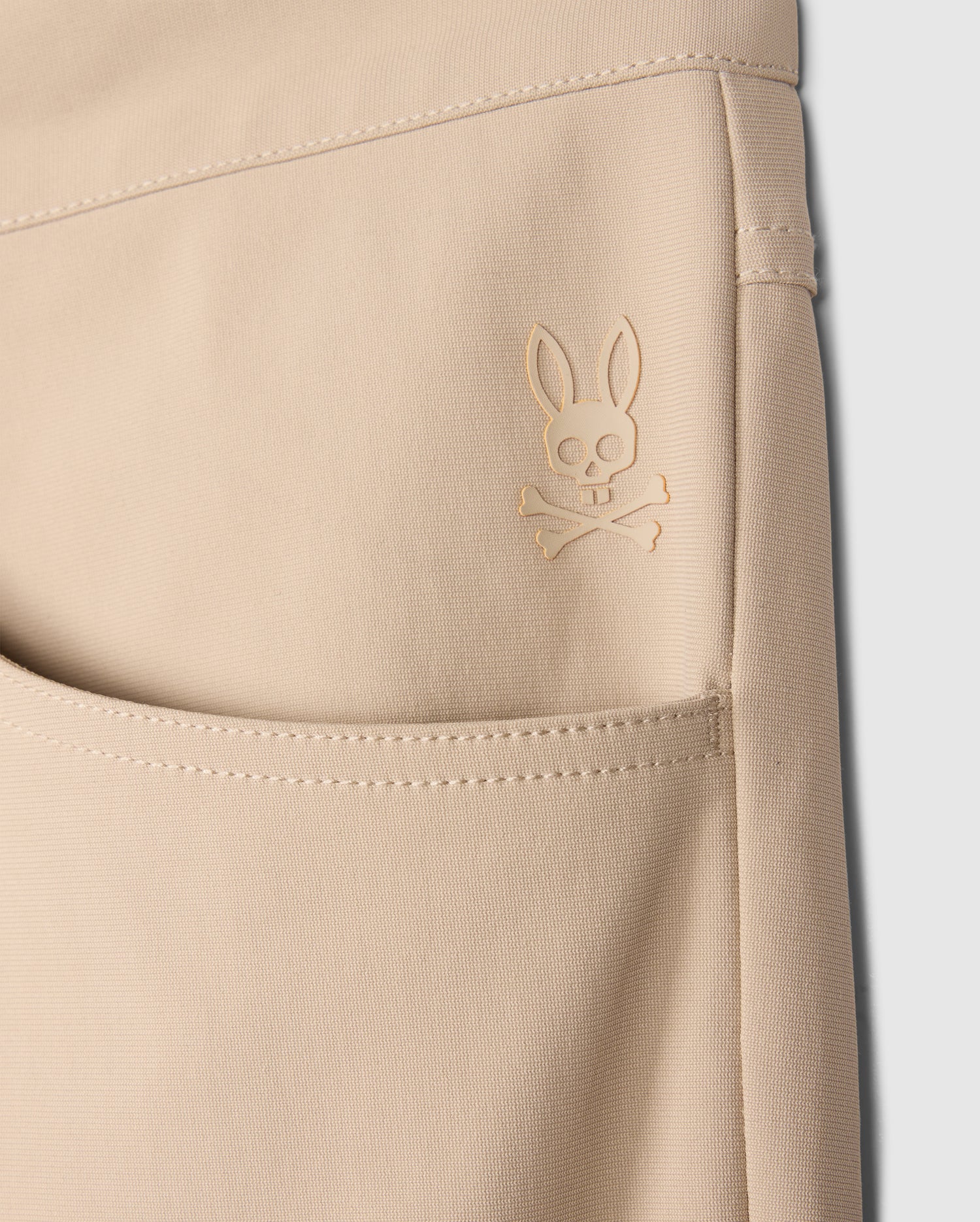 Close-up of a beige pocket on the MENS 10K STEP PANT - B6P788D200 by Psycho Bunny, featuring a raised emblem of a bunny skull and crossbones design. The stitching and fabric texture are visible, showcasing its four-way stretch material. The background is plain white, highlighting the details.