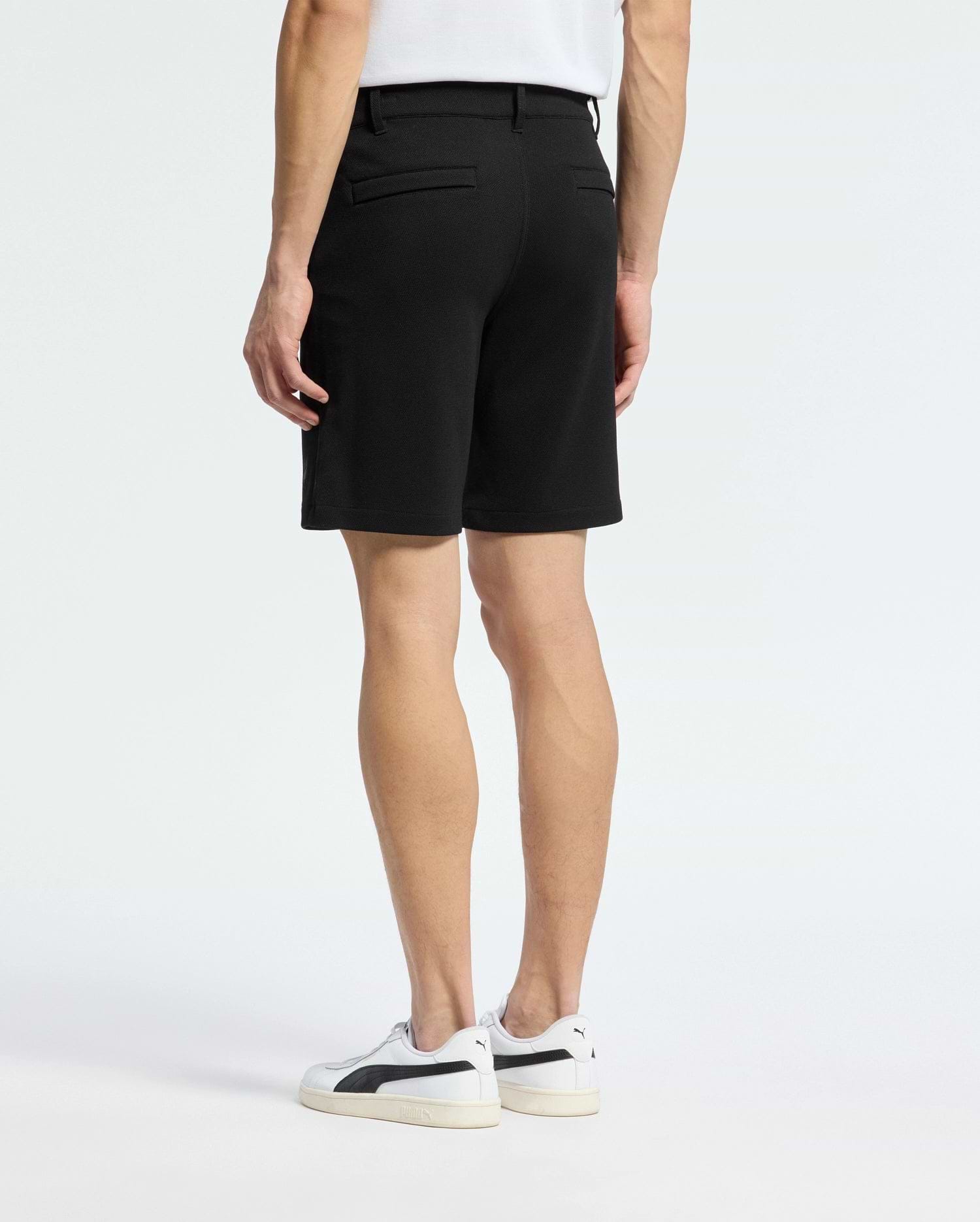 A person stands with their back to the camera, wearing a white shirt and the Psycho Bunny MENS SHIRO KNIT HONEYCOMB SHORT - B6R585F200. White sneakers with black accents complete the look against a plain background, showcasing a stylish transition for any setting.