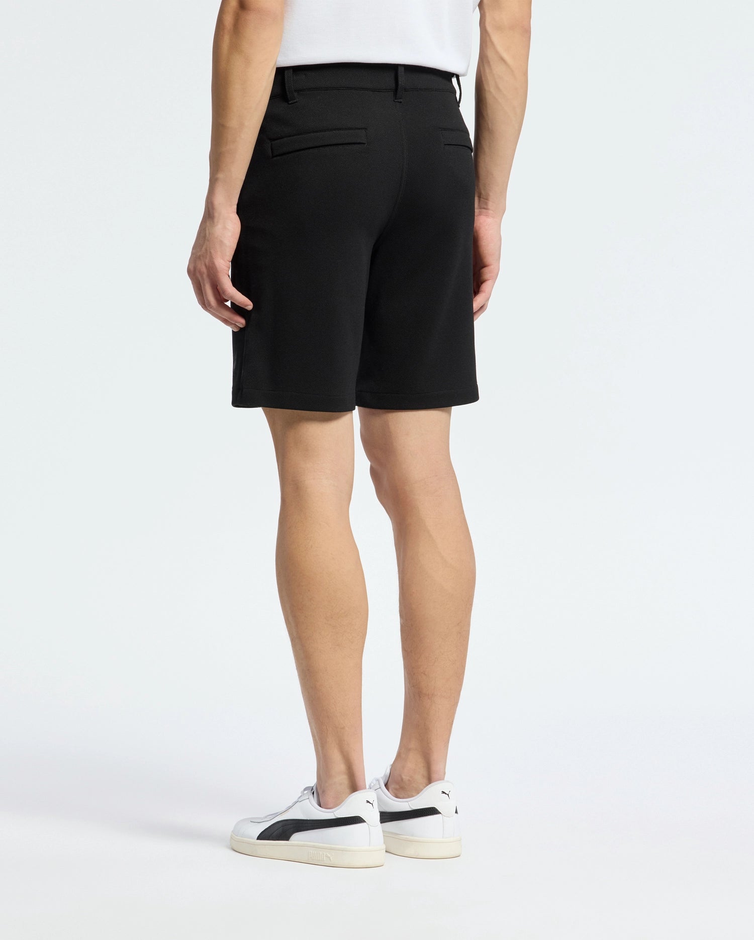 A person stands with their back to the camera, wearing a white shirt and the Psycho Bunny MENS SHIRO KNIT HONEYCOMB SHORT - B6R585F200. White sneakers with black accents complete the look against a plain background, showcasing a stylish transition for any setting.