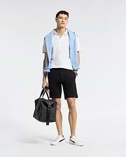 A person poses against a plain background, donning a white polo, versatile shorts, and white sneakers. Draped over their shoulders is Psycho Bunny's light blue MENS SHIRO KNIT HONEYCOMB SHORT. They hold a black bag in one hand, effortlessly transitioning from casual to chic.