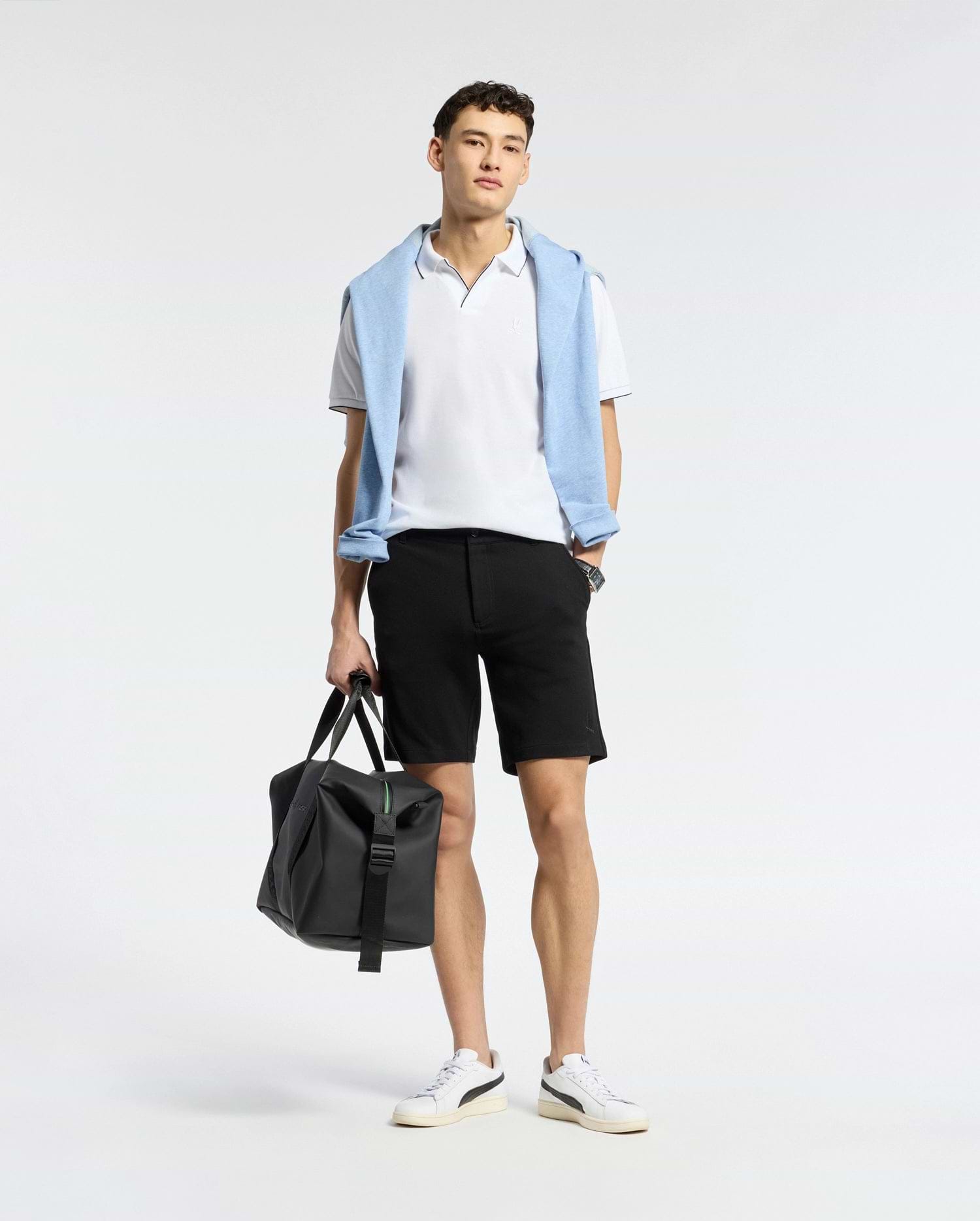 A person poses against a plain background, donning a white polo, versatile shorts, and white sneakers. Draped over their shoulders is Psycho Bunny's light blue MENS SHIRO KNIT HONEYCOMB SHORT. They hold a black bag in one hand, effortlessly transitioning from casual to chic.