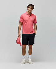 Coral pink pique polo with white tipped collar and cuffs, featuring a small embroidered blue and red Psycho Bunny skull logo on the left chest, paired with classic fit black Mens Maverick 10k Step Short.