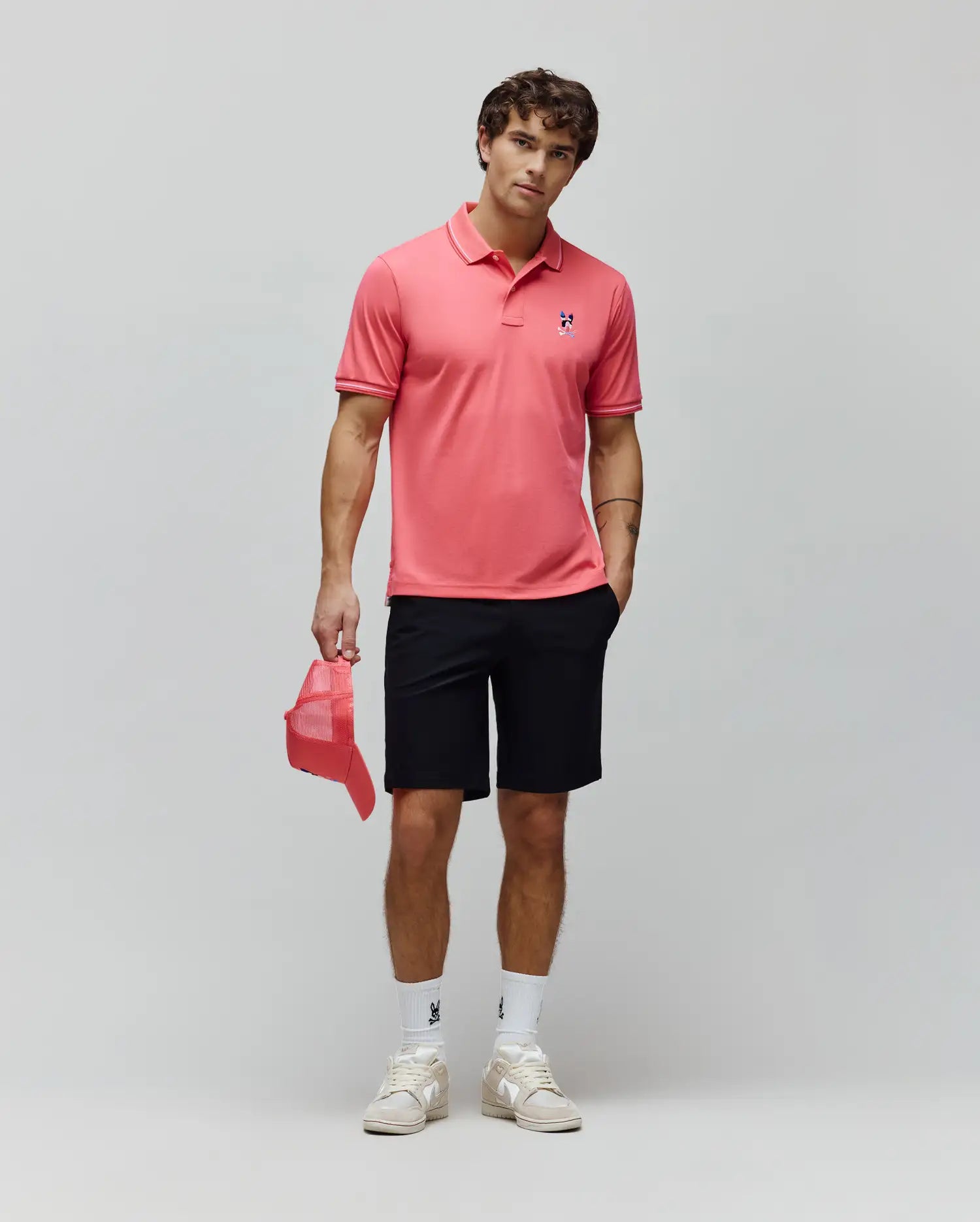 Coral pink pique polo with white tipped collar and cuffs, featuring a small embroidered blue and red Psycho Bunny skull logo on the left chest, paired with classic fit black Mens Maverick 10k Step Short.