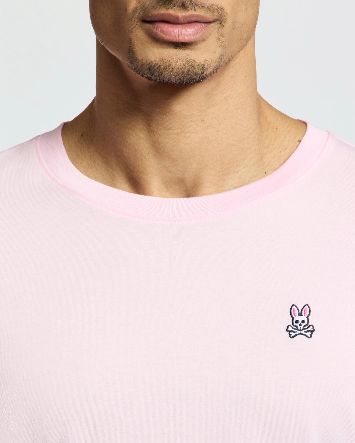 A person is shown from shoulders to just above the mouth, wearing a Psycho Bunny MENS CLASSIC CREW NECK TEE - B6U014CRPC in versatile light pink, made from soft Pima cotton with a classic crewneck and a small embroidered chest logo of a bunny face over crossed bones.