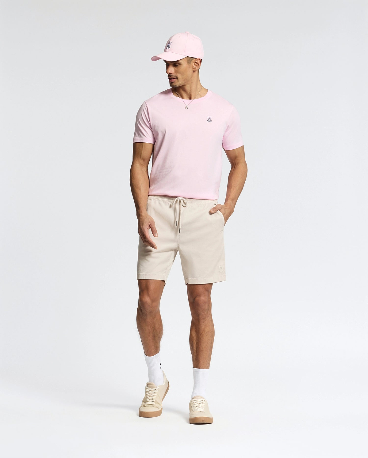 A man models the versatile pink Psycho Bunny MENS CLASSIC CREW NECK TEE - B6U014CRPC, paired with a matching pink cap, beige shorts, white socks, and beige sneakers. Made from soft pima cotton, he stands against a plain white background with his hands in his pockets, gazing to the side.