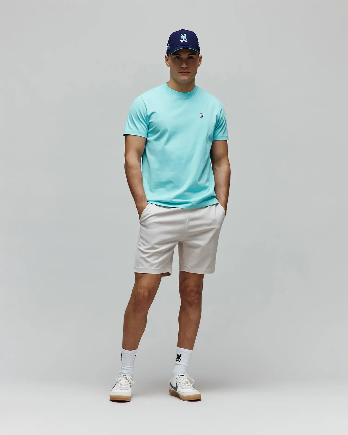 Mens Classic Crew Neck Tee in aqua jersey with small embroidered Psycho Bunny logo, paired with light beige shorts, a navy cap with light blue embroidered logo, and white socks with a black bunny skull.