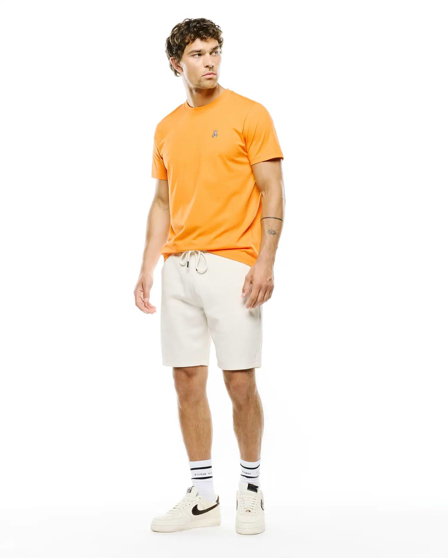 Mens Classic Crew Neck Tee in vibrant orange, crafted from smooth jersey fabric with a classic fit, featuring a small embroidered Psycho Bunny skull logo on the left chest. Cream french terry shorts complete the look.