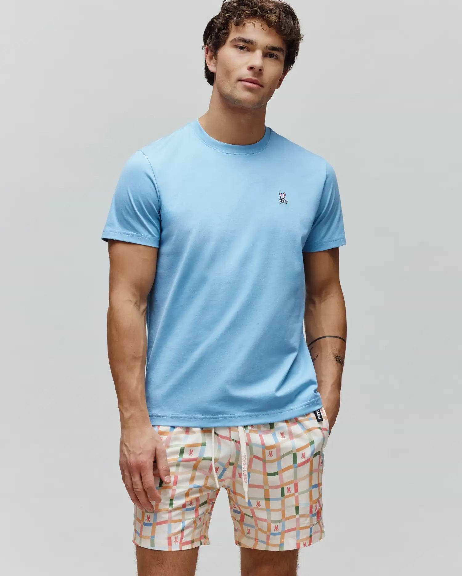 Mens Classic Crew Neck Tee in light blue jersey, with a small embroidered Psycho Bunny skull logo on the left chest. Paired with cream multi-color plaid shorts featuring repeating bunny skull patterns.