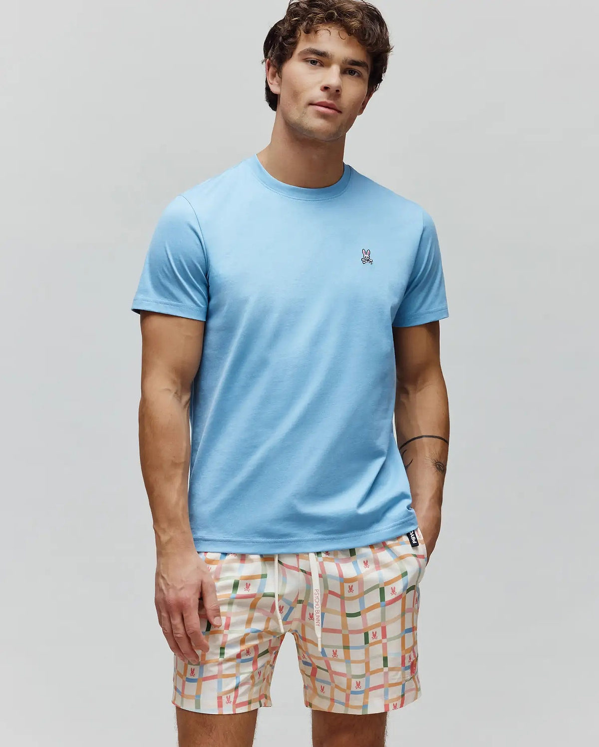 Mens Classic Crew Neck Tee in light blue jersey, with a small embroidered Psycho Bunny skull logo on the left chest. Paired with cream multi-color plaid shorts featuring repeating bunny skull patterns.