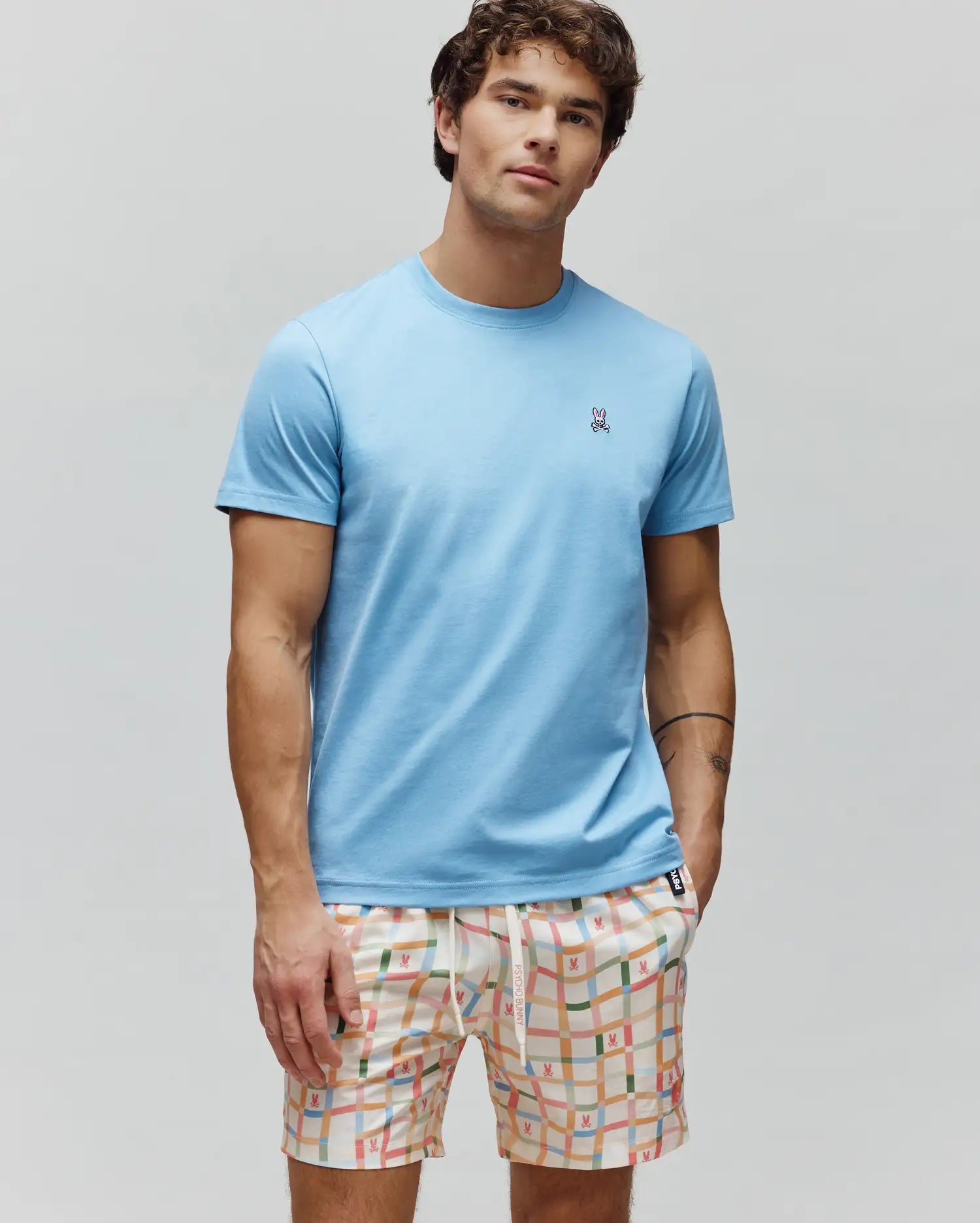 Mens Classic Crew Neck Tee in light blue jersey, with a small embroidered Psycho Bunny skull logo on the left chest. Paired with cream multi-color plaid shorts featuring repeating bunny skull patterns.