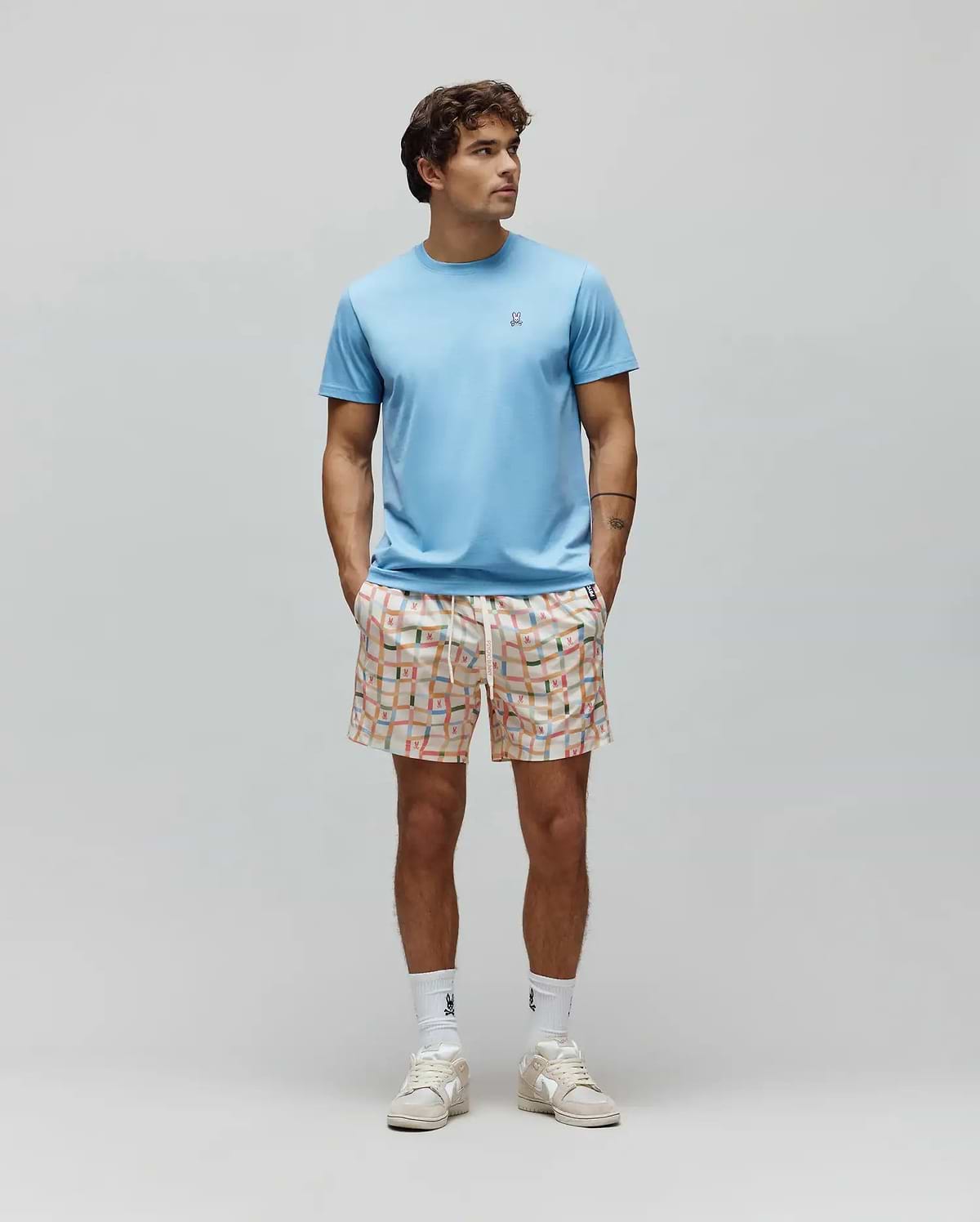 Light blue Mens Classic Crew Neck Tee, classic fit jersey, with small embroidered Psycho Bunny skull logo. Paired with cream relaxed fit shorts, vibrant multi-color grid pattern, and integrated bunny skull logos.