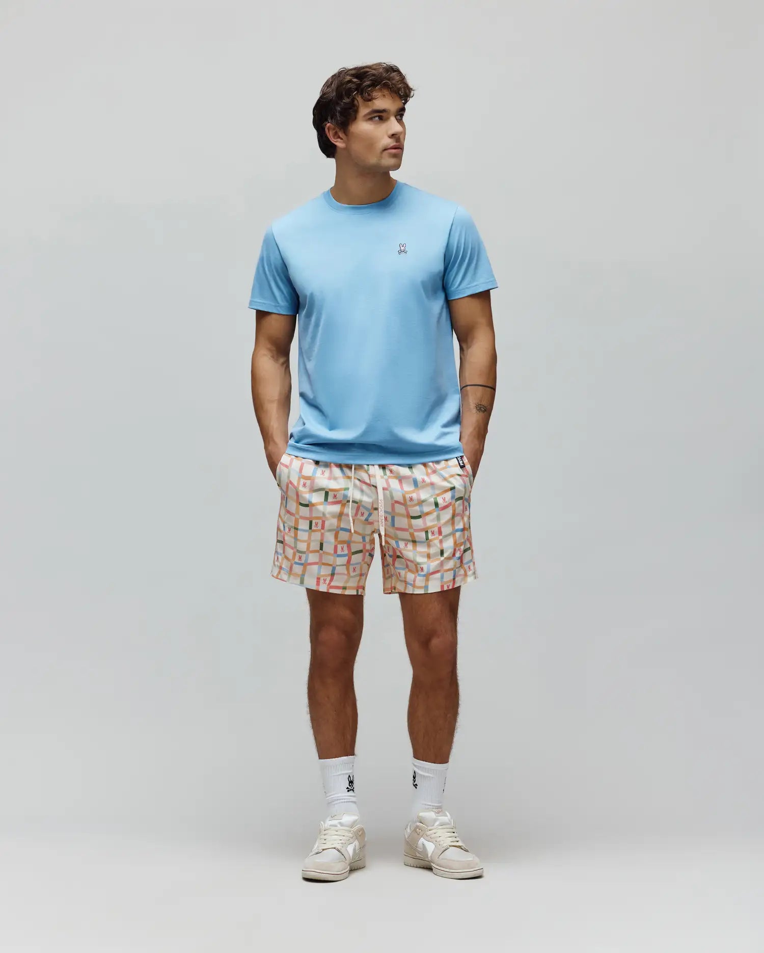 Light blue Mens Classic Crew Neck Tee, classic fit jersey, with small embroidered Psycho Bunny skull logo. Paired with cream relaxed fit shorts, vibrant multi-color grid pattern, and integrated bunny skull logos.